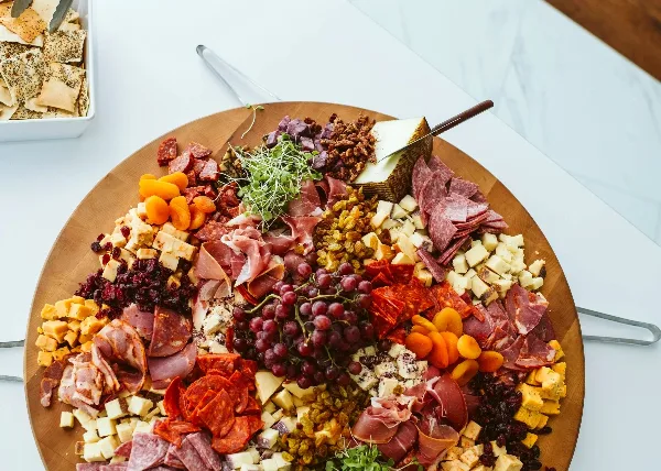 A large round wooden board filled with assorted cured meats, cheeses, grapes, dried fruits, and garnishes, presented on a white table.