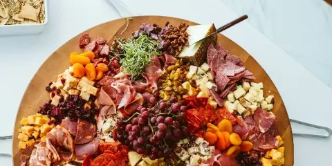 A large round wooden board filled with assorted cured meats, cheeses, grapes, dried fruits, and garnishes, presented on a white table.