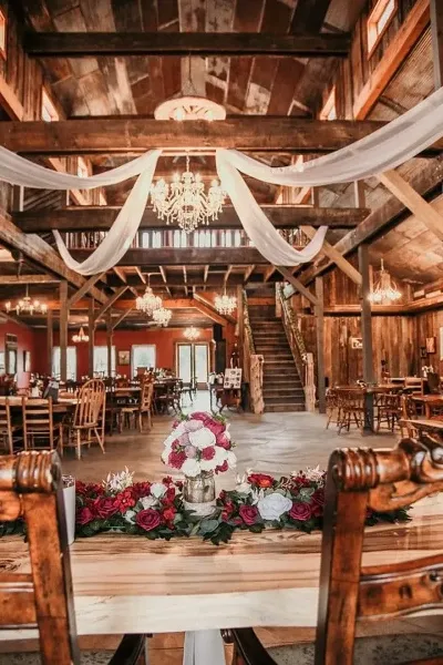 Interior view of a rustic wedding venue featuring wooden beams, crystal chandeliers, and a long table decorated with a floral centerpiece.