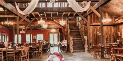 Interior view of a rustic wedding venue featuring wooden beams, crystal chandeliers, and a long table decorated with a floral centerpiece.