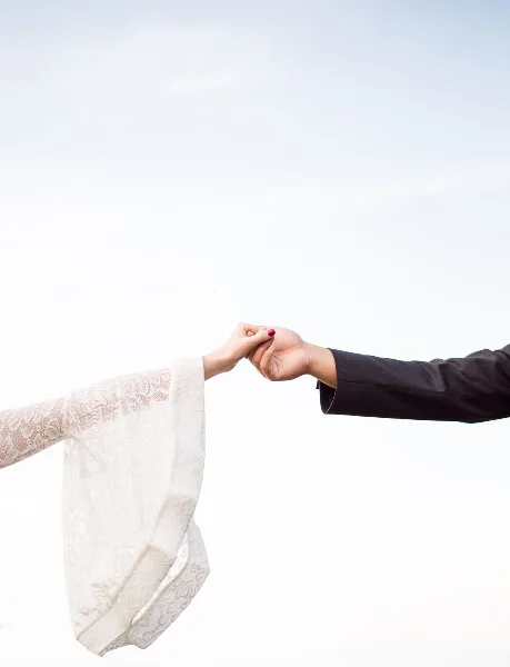 A bride in a white lace dress and a groom in a dark suit holding hands against a bright sky.