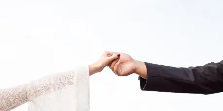 A bride in a white lace dress and a groom in a dark suit holding hands against a bright sky.