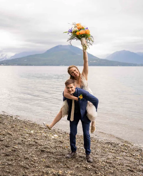 A bride and groom posing playfully on a rocky shoreline with a lake and mountains in the background.