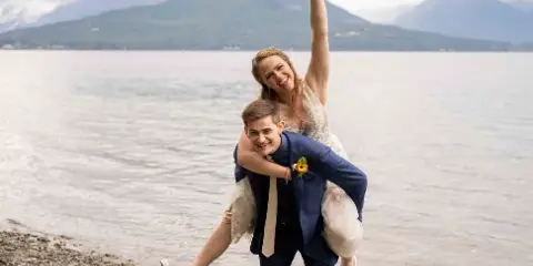 A bride and groom posing playfully on a rocky shoreline with a lake and mountains in the background.