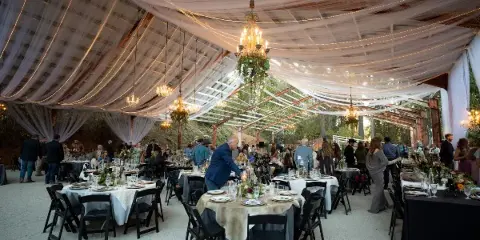 Wide shot of a wedding reception venue featuring a glass roof, draped fabric, chandeliers, and set dining tables.