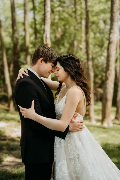 A bride and groom embracing in a wooded area, with the bride wearing a white lace dress and the groom in a black suit.
