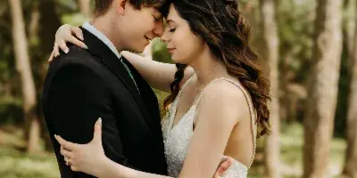 A bride and groom embracing in a wooded area, with the bride wearing a white lace dress and the groom in a black suit.