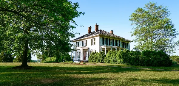 Exterior view of a white two-story historic manor house with black shutters, set on a green lawn with mature trees.