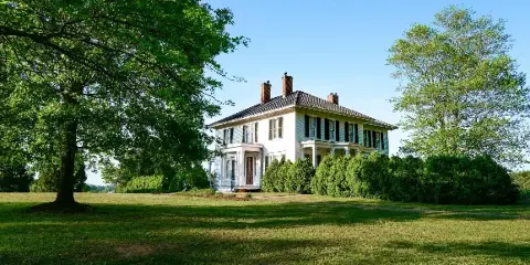 Exterior view of a white two-story historic manor house with black shutters, set on a green lawn with mature trees.