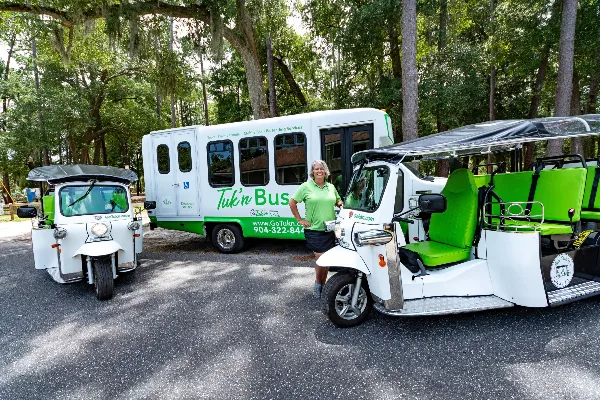 A staff member in a green polo shirt stands between a white tuk-tuk and a white shuttle bus labeled 'Tuk'n Bus' in a wooded area.