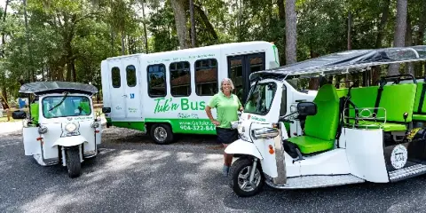 A staff member in a green polo shirt stands between a white tuk-tuk and a white shuttle bus labeled 'Tuk'n Bus' in a wooded area.