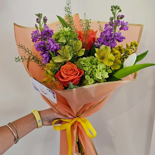 A hand holding a colorful floral bouquet wrapped in peach paper and tied with a yellow ribbon.