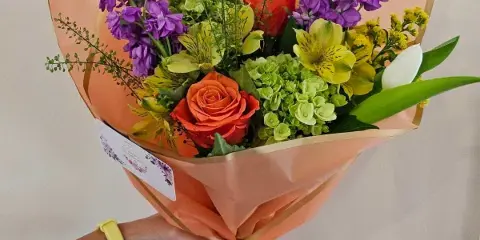 A hand holding a colorful floral bouquet wrapped in peach paper and tied with a yellow ribbon.