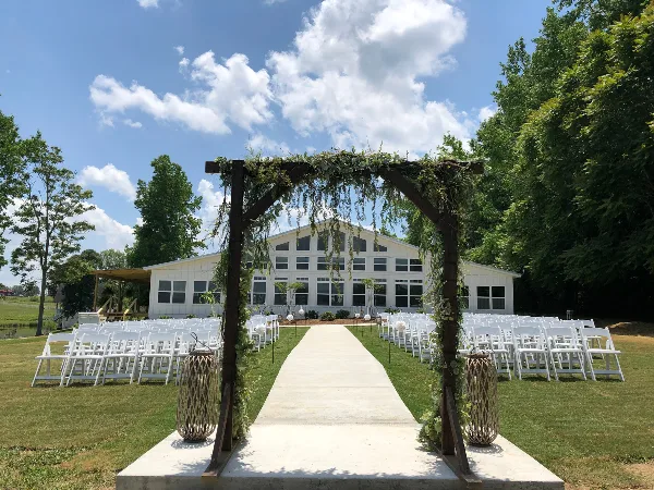 Outdoor wedding ceremony setup with a wooden arch, white chairs, and a large white venue building in the background.