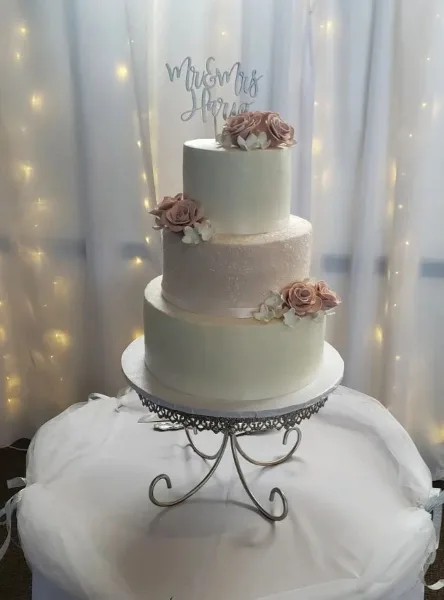 A three-tiered white wedding cake with pink floral decorations and a Mr & Mrs topper on a metal stand.