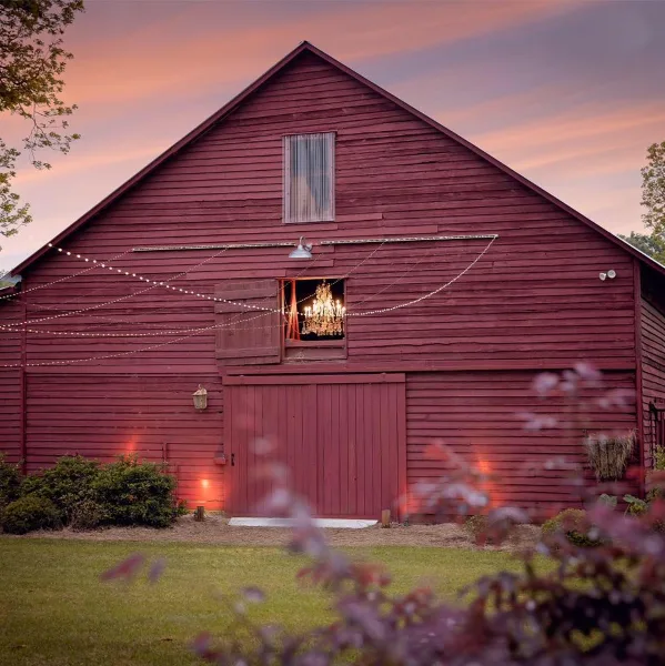 Exterior view of a red wooden barn event venue at dusk with string lights and a chandelier visible in the window.