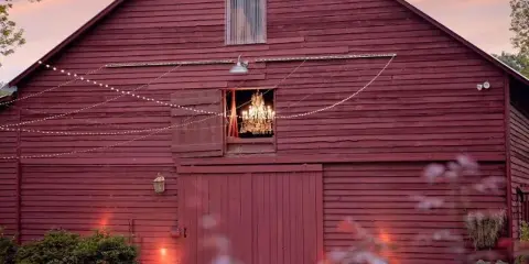 Exterior view of a red wooden barn event venue at dusk with string lights and a chandelier visible in the window.