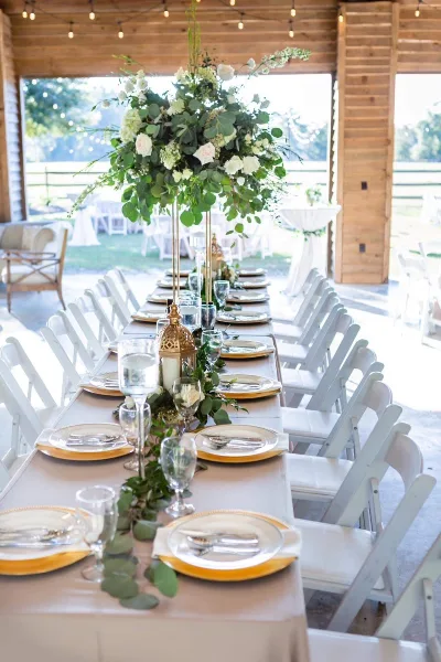 A long wedding reception table set with white and gold place settings, floral centerpieces, and white chairs inside a rustic wooden barn venue.