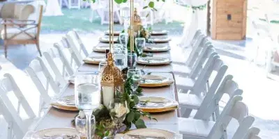 A long wedding reception table set with white and gold place settings, floral centerpieces, and white chairs inside a rustic wooden barn venue.