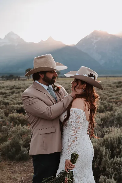A bride and groom in cowboy hats and formal wear embracing in a sagebrush field with mountains in the background.