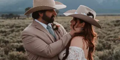 A bride and groom in cowboy hats and formal wear embracing in a sagebrush field with mountains in the background.