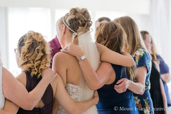 A bride in a white lace wedding dress and veil hugs a group of women in a bright indoor setting.