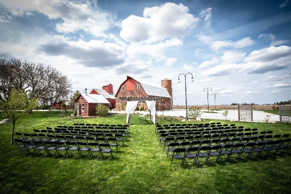 Outdoor wedding ceremony setup with rows of black chairs on a lawn in front of a red brick barn and silo under a cloudy sky.