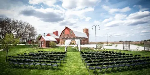 Outdoor wedding ceremony setup with rows of black chairs on a lawn in front of a red brick barn and silo under a cloudy sky.