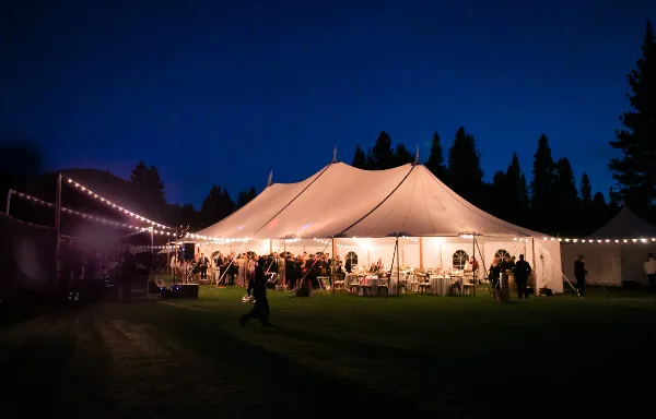 A large white event tent illuminated by string lights at dusk, set up on a grassy field with tables and guests visible inside.