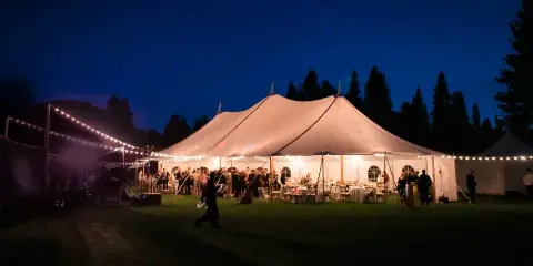 A large white event tent illuminated by string lights at dusk, set up on a grassy field with tables and guests visible inside.