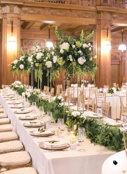 A formal event table setting with white linens, gold-rimmed plates, and tall floral centerpieces featuring white flowers and greenery.