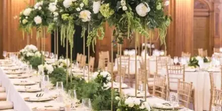 A formal event table setting with white linens, gold-rimmed plates, and tall floral centerpieces featuring white flowers and greenery.