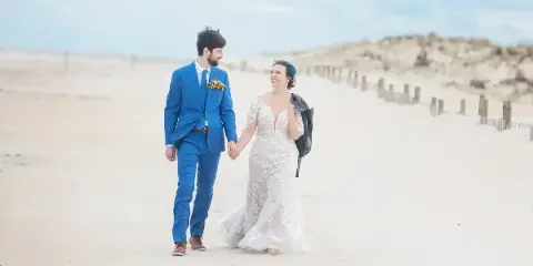A bride and groom holding hands and walking on a sandy beach.