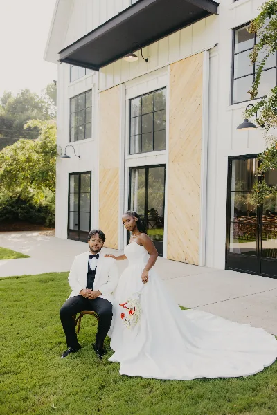 A bride in a white gown and a groom in a white tuxedo jacket posing together on a lawn in front of a white barn-style building.
