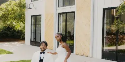 A bride in a white gown and a groom in a white tuxedo jacket posing together on a lawn in front of a white barn-style building.