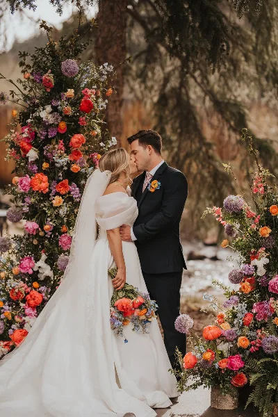 A bride and groom kissing in front of a large, colorful floral arch and holding a bouquet.