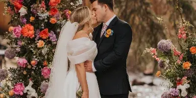 A bride and groom kissing in front of a large, colorful floral arch and holding a bouquet.