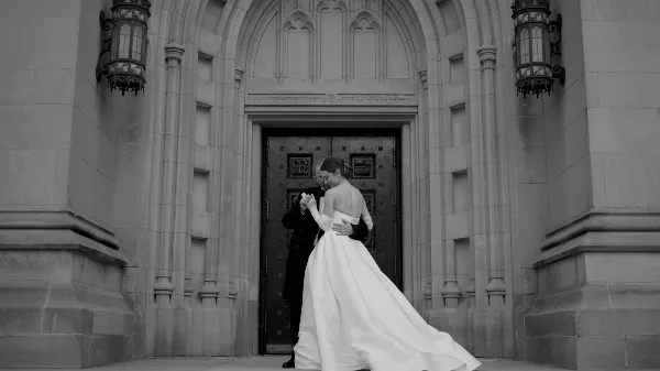 A black and white cinematic photograph of a bride and groom embracing in front of a grand stone archway.