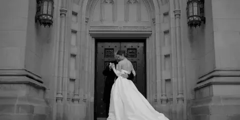 A black and white cinematic photograph of a bride and groom embracing in front of a grand stone archway.