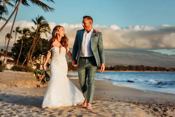 A bride and groom walking hand-in-hand on a sandy beach with palm trees and the ocean in the background.