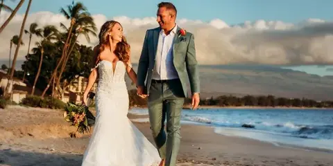 A bride and groom walking hand-in-hand on a sandy beach with palm trees and the ocean in the background.