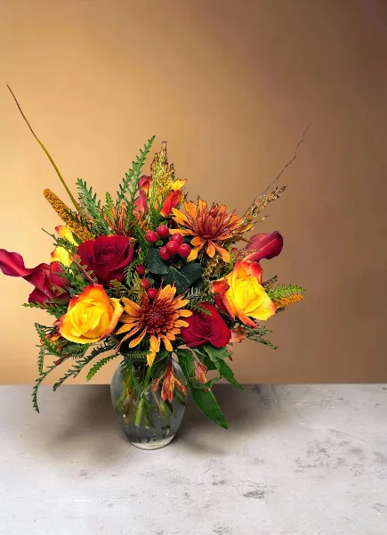 A vibrant floral arrangement with red and yellow roses, orange chrysanthemums, and greenery in a clear glass vase.