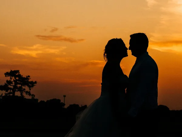 Silhouette of a bride and groom facing each other against a vibrant orange sunset sky.