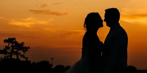 Silhouette of a bride and groom facing each other against a vibrant orange sunset sky.