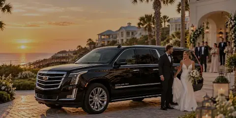 A black Cadillac Escalade ESV parked on a cobblestone driveway at sunset, with a groom assisting a bride out of the vehicle while a wedding party stands in the background.