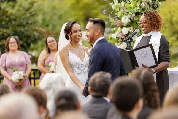 A wedding officiant wearing a clerical collar and holding a branded folder officiates a couple during an outdoor ceremony.