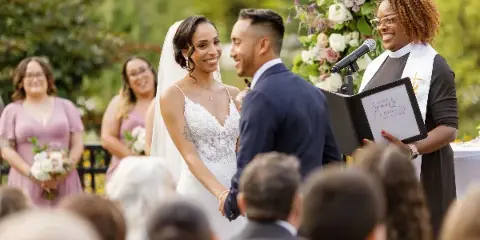 A wedding officiant wearing a clerical collar and holding a branded folder officiates a couple during an outdoor ceremony.