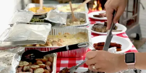 A catering table with various food trays and people serving themselves.