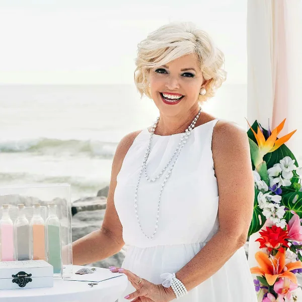 A smiling woman in a white dress standing next to a table with a ring box and wedding flowers.
