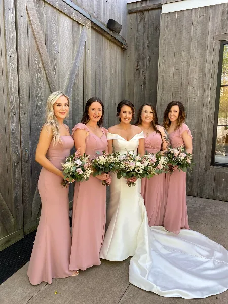 Bride and bridesmaids holding custom floral bouquets in front of a wooden barn wall.
